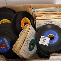 Large box filled with approximately 50 45 RPM vinyl records, some with colorful labels and protective sleeve covers.