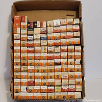 Photo of a large assortment of vacuum tubes in mostly orange and white GE branded boxes with some RCA and other brands scattered throughout, arranged in rows inside a cardboard container.