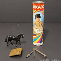 Photo of the lot showing the Mikado game tube, black metal horse figurine, brass Caterpillar belt buckle, and antique pipe tool arranged together.