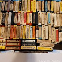 Overhead image showing a large collection of assorted 8-track tapes arranged in rows inside a box; cases have varied colors and visible wear.