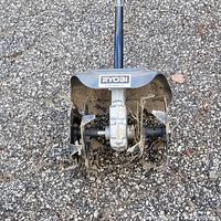 Top view of Ryobi rotor tiller base showing soil tilling tines and Ryobi branding on metal housing and shaft.