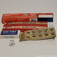 Photo showing two Trico Rainbow wiper blades in original packaging, tube of Permatex Super Weatherstrip Adhesive, box of Spectro miniature ampoules, and assorted automotive bulbs.