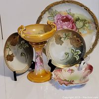 Photo showing five vintage porcelain dishes arranged: two small R & S Germany plates with gold rose motifs, a large plate with pink roses and gold rim, a rose-shaped candy dish, and a brown pedestal candy dish with two handles.