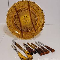 Amber glass Tiara platter with carved pattern on rim and center starburst, arranged upright with flatware in front.
