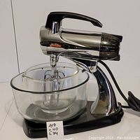 Front-side view showing Sunbeam mixer with large glass bowl fitting around the beaters attached to mixer head. Mixer has chrome finish and black base.