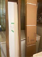 Two long cardboard boxes containing unassembled shelving units in box, one opened showing white panels and instruction sheet attached to inner panel.