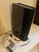 Oreck air purifier standing upright on a white surface with power cord coiled nearby and user guide placed beside it.