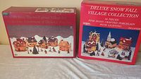Two unopened boxed Christmas village sets displayed side by side, one red with a photo of the village pieces on the front, the other red with 'Deluxe Snow Fall Village Collection' labeling.