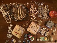 Full view of jewelry lot spread out on wooden surface showing multiple necklaces, clip on earrings, brooches, bracelet, locket, small decorative items, and music box parts