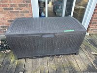 Closed deck box with textured dark brown plastic weave pattern on a wooden deck in front of a brick wall.