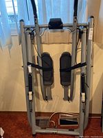 Front view of the Gazelle Freestyle Crosstrainer showing silver tubular frame, black foot pedals, and a small central display unit. Unit appears folded upright against a wall.