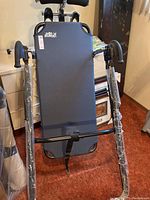 Front view of folding inversion table showing blue padded backrest, black handles, and protective plastic on legs.