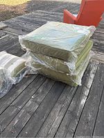 Four olive green outdoor cushions stacked on a wooden deck, each wrapped in clear plastic, showing their square shape and thickness.