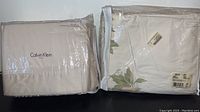 Calvin Klein queen sheet set and Bella queen duvet ensemble side by side in packaging, showing brand labels and packaging condition.