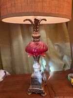 Photo showing full table lamp with beige drum textured lampshade, red swirled glass base and ornate silver-toned metal stand on wooden surface with curtain background.