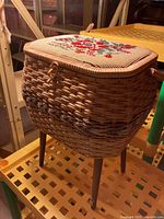 Side view of vintage wicker sewing basket on wooden legs with floral needlepoint lid.