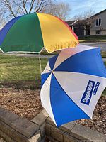 Two sun umbrellas shown outdoors on a clear day. One umbrella is multi-coloured with red, yellow, green, and blue panels. The second umbrella is blue and white with a 'Coppertone' logo visible on the blue panel.