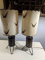 Pair of mid-century modern table lamps with hairpin legs and perforated cylindrical metal bases, cream-colored shades with dark patterns, photographed on a white surface.