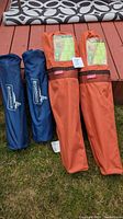 Four camping items laid on grass: Two blue carrying bags for chairs with Promarines logos and two orange carrying bags for side tables with Coleman logo.