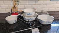 Four white ceramic Corningware casseroles with blue cornflower design, two glass lids, two metal BBQ forks with black handles, and one red plastic BBQ spatula on black countertop