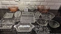 Overview photo of the full lot including varied glass serving pieces such as bowls, trays, casseroles, and a pitcher, showing clear and amber/pink hues with intricate pressed patterns.