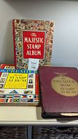 Photo of three stamp albums: The Majestic Stamp Album with colorful cover and two Premier Model Premier albums, one maroon and one blue, showing wear on covers and edges.