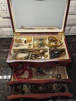 Open mahogany-finish jewelry box showing mirror, top compartment and two drawers filled with assorted jewelry