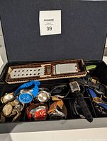 Box containing multiple assorted wristwatches with varied bands and faces, including metal, leather, and fabric bands in colors such as blue, black, and brown.
