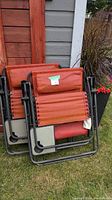 Two folded zero gravity chairs with orange fabric seat and moveable head cushions leaning against a wooden fence outside.