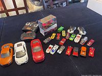 Full view of all assorted cars including three larger sports cars and smaller vintage, racing, and model cars, with Tesla Model 3 in lucite box prominently displayed.