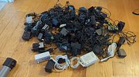 Group photo showing a large pile of assorted AC adapters on a wooden floor, various sizes, colors mostly black with a few white adapters, tangled cords.
