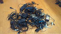 Photo showing a large pile of various laptop chargers and power adapters tangled with cables on a wood floor.