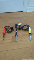 Three soldering irons with colored handles (orange, black, white/yellow) and attached cords laid on wooden floor