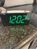 Front and top view of the black LED digital alarm clock displaying 12:02 in green digits, showing two round buttons on top and glossy reflective surface.