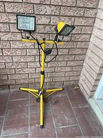 Twin head halogen work light mounted on yellow telescoping tripod stand, showing front and side views with cord and switches.