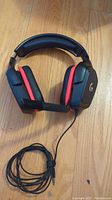 Front view of Logitech G332 wired gaming headset with black and red ear cushions on wood floor, cable coiled neatly.