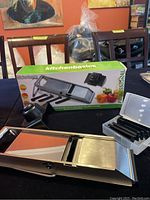 Wide angle photo showing the mandoline slicer with its box, blades case, and mesh tray on table.