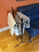 Black metal blanket holder with two blankets hanging, showing structure and decorative side scrolls near a blue leather couch and orange wall.