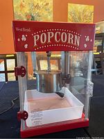 Front view of West Bend popcorn maker showing brand logo, red top, clear sides, silver popping kettle, and white tray inside