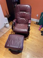 Front view of the wine-colored leather reclining arm chair and matching ottoman on a hardwood floor with wall and other household items in background.