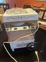 Front view of vintage style Nostalgia snow cone maker machine with white and gold trim, showing clear ice container and electric cord.