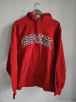 Front view of a red Supreme hoodie with embroidered 'SUPREME' arc logo in black and white hearts.