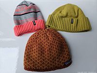 Three Patagonia winter hats displayed against a white background: a grey hat with coral and navy stripes, a green ribbed knit hat, and a multicolor knit hat in orange/yellow tones