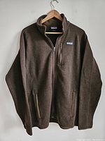 Front view of men's brown Patagonia fleece jacket hanging on wooden hanger, showing full front with collar, zippered pockets, and Patagonia logo patch on left chest.
