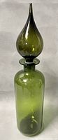 Full view of the olive green blown glass genie bottle with tall curved stopper on gray background.