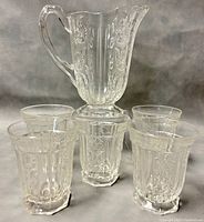 Photo showing the clear cut glass pitcher with floral patterns and five matching footed juice glasses arranged in front.