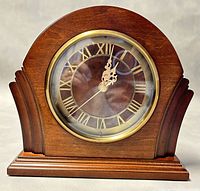 Front view of Bombay wooden mantle clock showing brown wood arched design and brass accented round clock face with Roman numerals and ornate hands