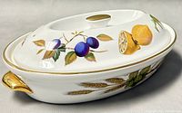 Oval covered baking dish with white ceramic base, decorated with painted fruit (lemons, plums) and gold trim on handles and lid knob.