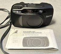 Front view of black Minolta 115EX Supreme Freedom Zoom camera with lens cover closed, showing brand, model name, and passive AF labeling. Wrist strap visible.