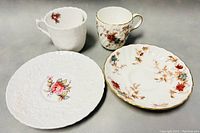 Photo showing two cups and two saucers, one white cup embossed with subtle floral pattern and a smaller floral cup with delicate gold trim, plus matching saucers